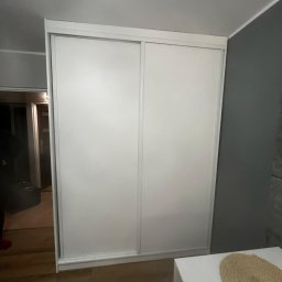 Daniel Jesionkowski - Biała szafa wnękowa z przesuwnymi drzwiami, zamontowana w mieszkaniu w Mosinie, widziana z boku, z fragmentem stołu w kadrze.