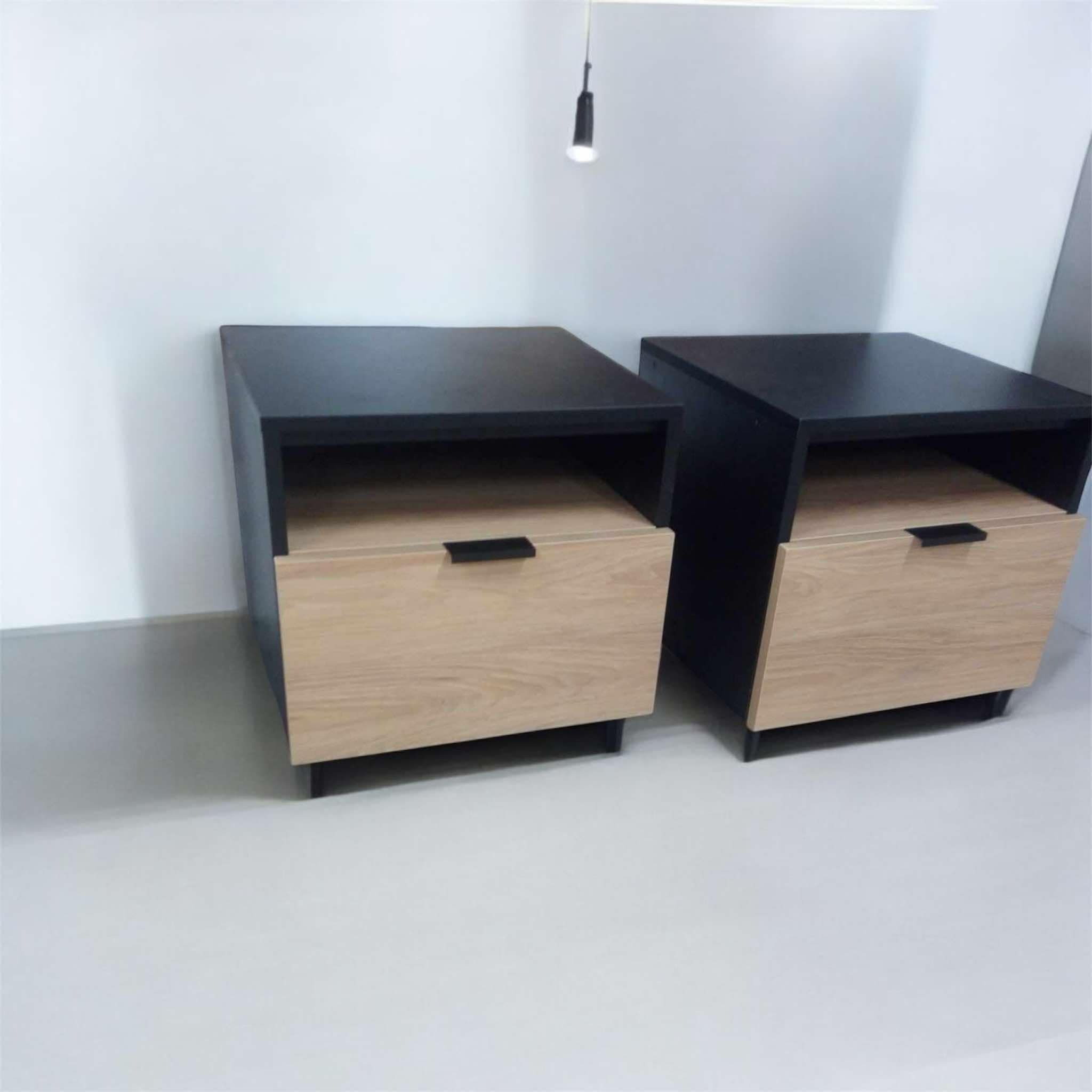 Dwie minimalistyczne szafki nocne z jasnego drewna i czarnymi blatami, ustawione na jasnej podłodze pod wiszącą lampą.