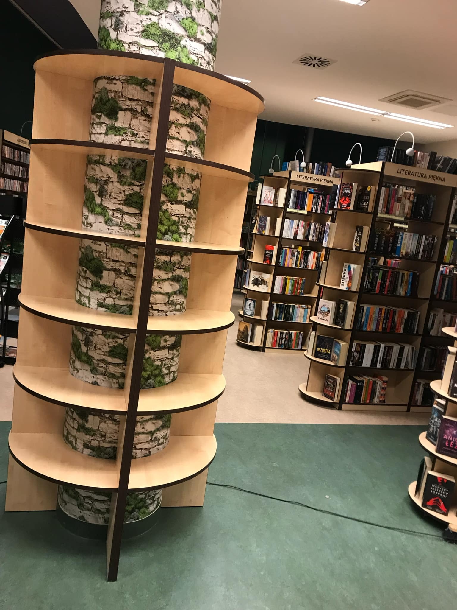 Nietypowy regał na książki z półkami w kształcie okręgów, z cylindrycznymi elementami dekoracyjnymi imitującymi mur z mchem, w tle inne regały z książkami w księgarni.