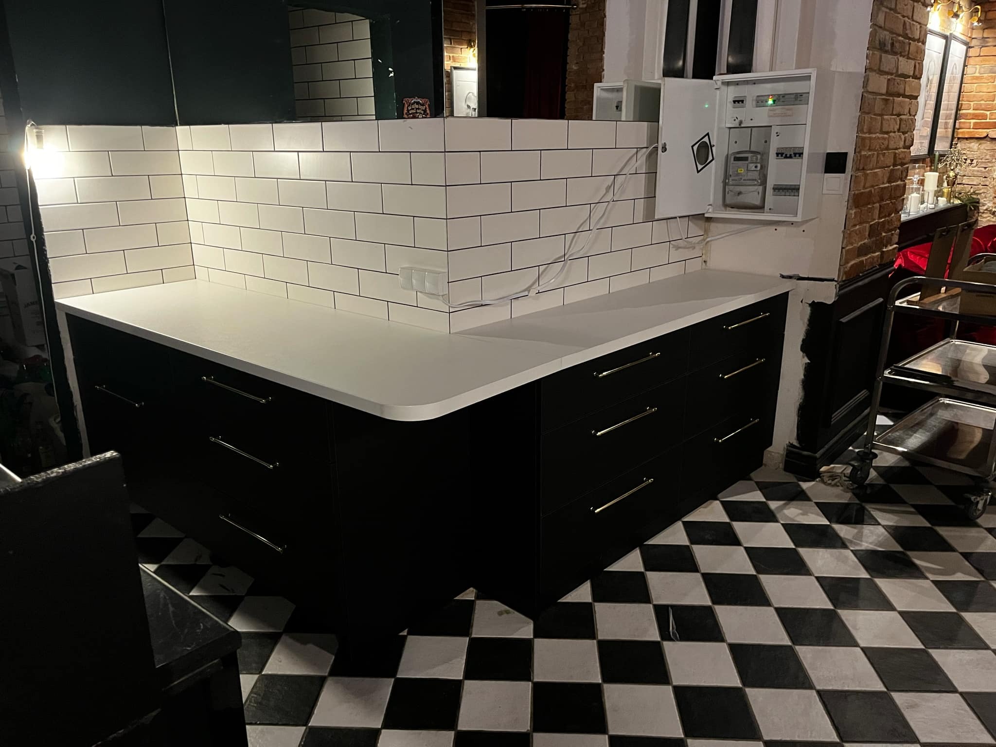 Czarne szafki z białym blatem i białymi kafelkami na ścianie, uzupełnione skrzynką elektryczną, na podłodze czarno-biała szachownica.