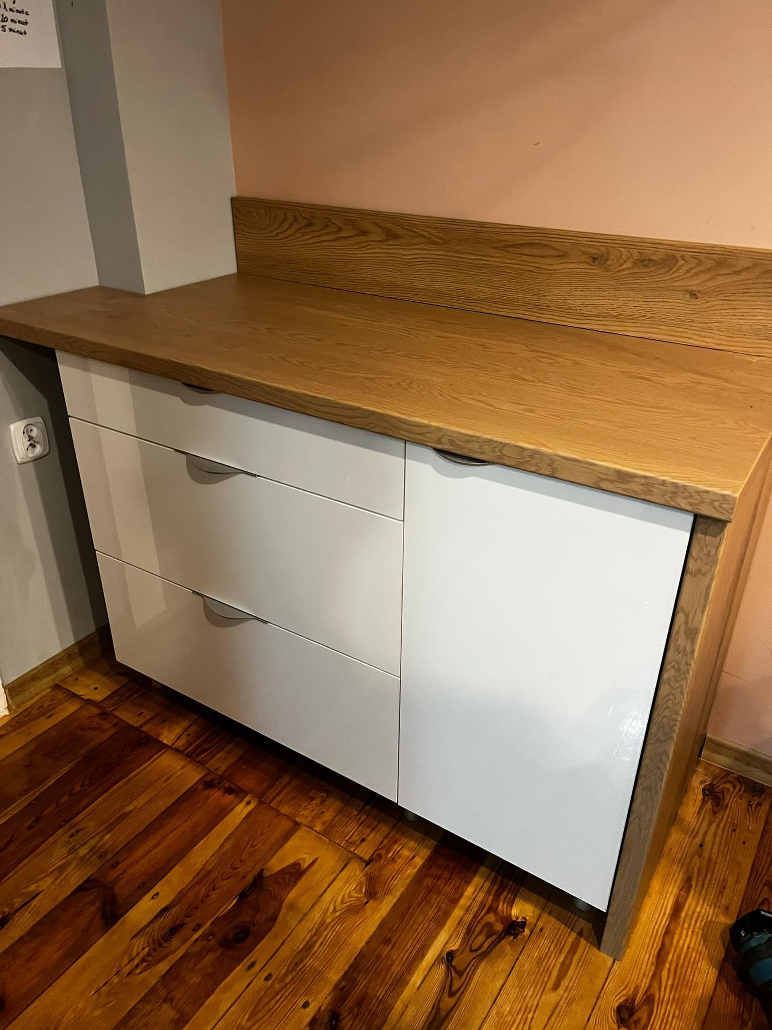 Nowoczesna komoda z trzema białymi szufladami i blatem z jasnego drewna, stojąca na drewnianej podłodze, widoczny fragment ściany w dwóch odcieniach.