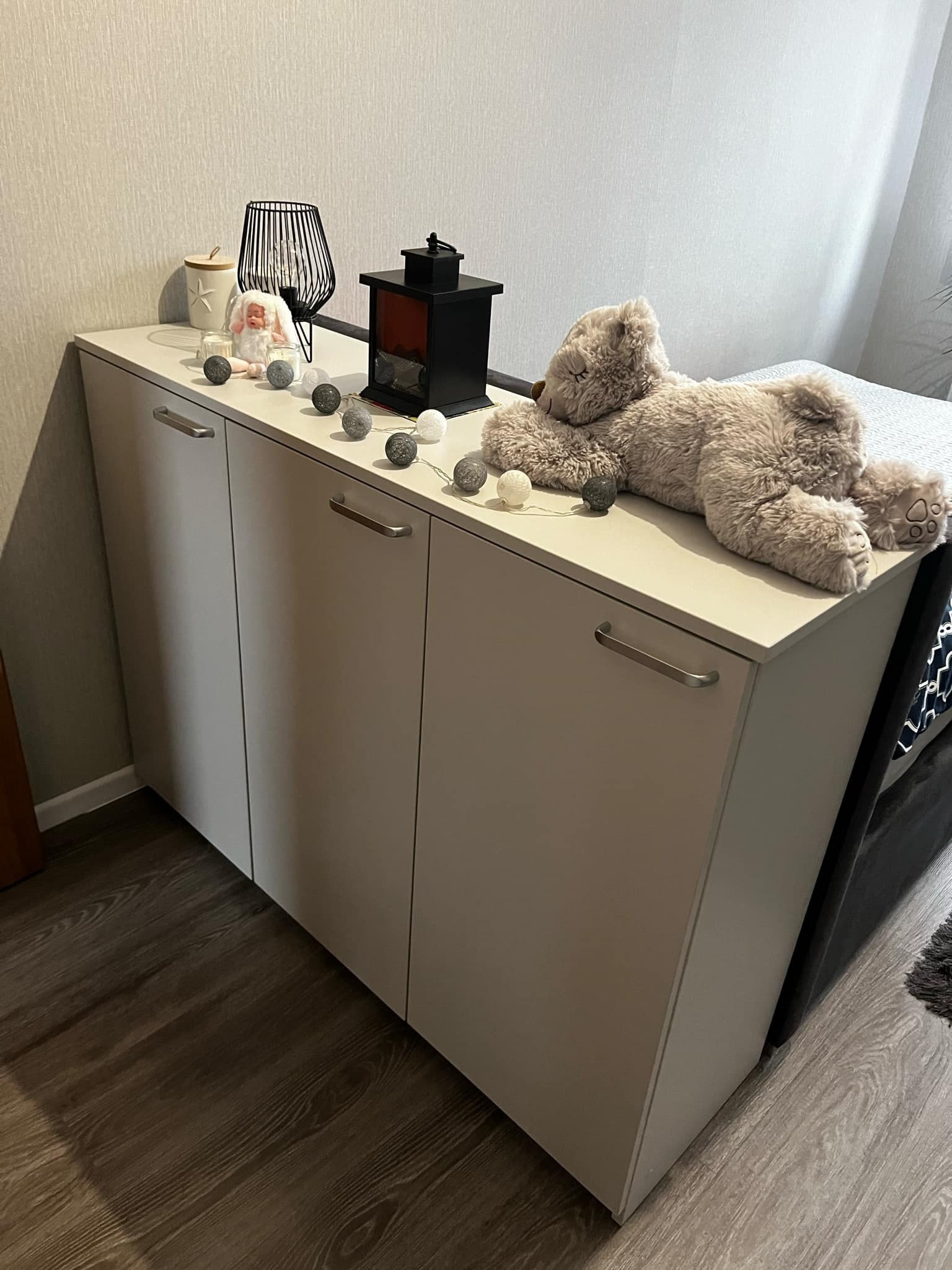 Trzydrzwiowa szafka z szarymi frontami i podłużnymi uchwytami, udekorowana lampkami cotton balls, lampionem i pluszowym misiem leżącym na blacie, częściowo zasłaniającym łóżko.