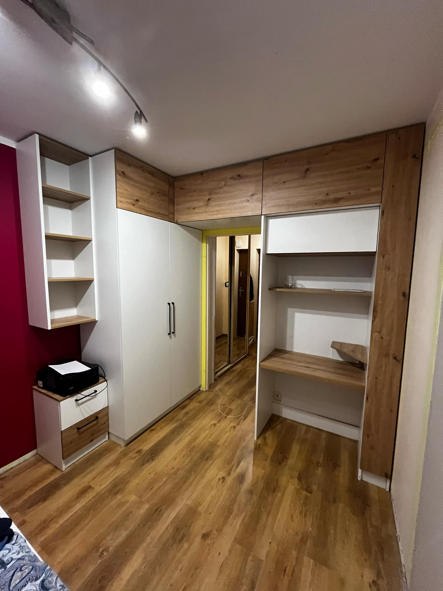 Biała zabudowa meblowa z elementami drewnopodobnymi w pokoju, obejmująca szafę z czarnymi uchwytami, regał z półkami i biurko z półkami, oświetlona lampą sufitową na szynie.