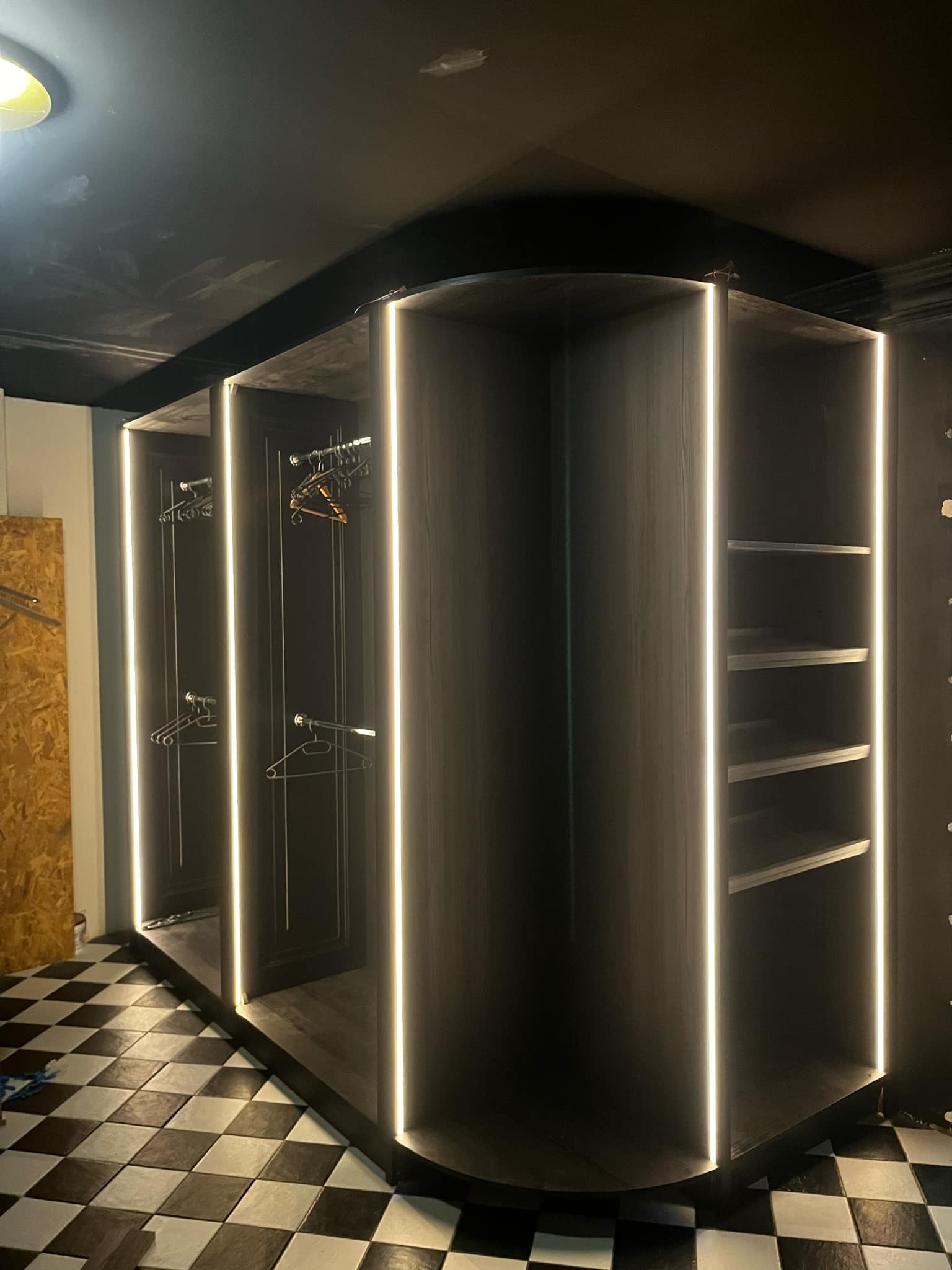 Czarna, narożna garderoba z oświetleniem LED, widoczne półki i drążki na wieszaki, na podłodze czarno-biała szachownica.