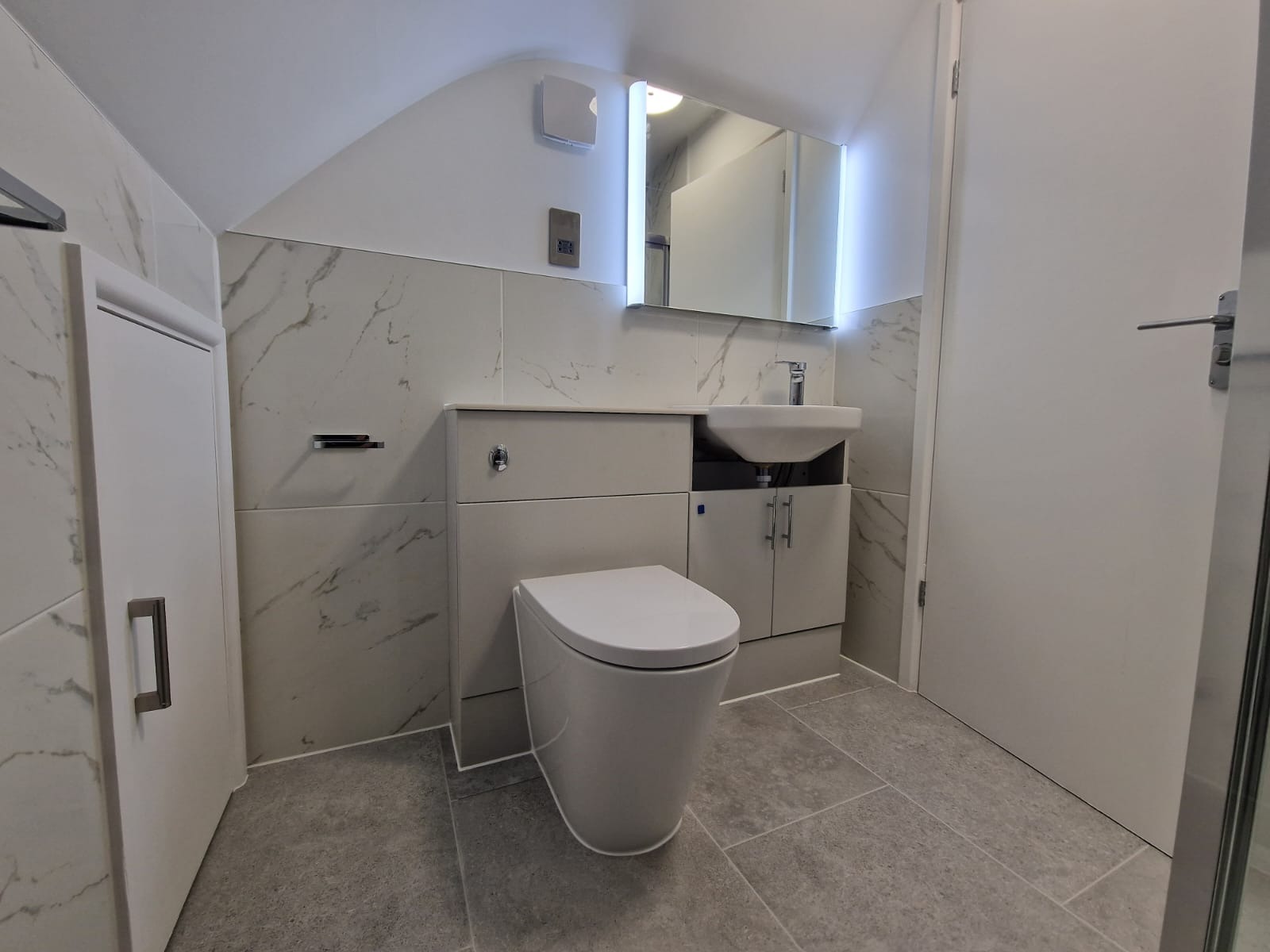 Nowoczesna łazienka z podwieszaną toaletą, umywalką wbudowaną w szafkę i lustrem z podświetleniem LED, wykończona płytkami imitującymi marmur.