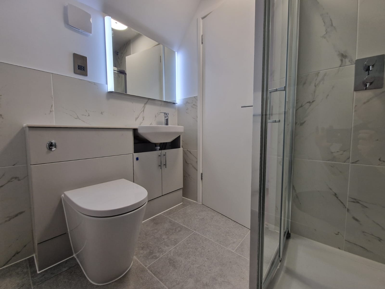 Nowoczesna łazienka z marmurowymi płytkami, podświetlanym lustrem, wiszącą toaletą i kabiną prysznicową ze szklanymi drzwiami.