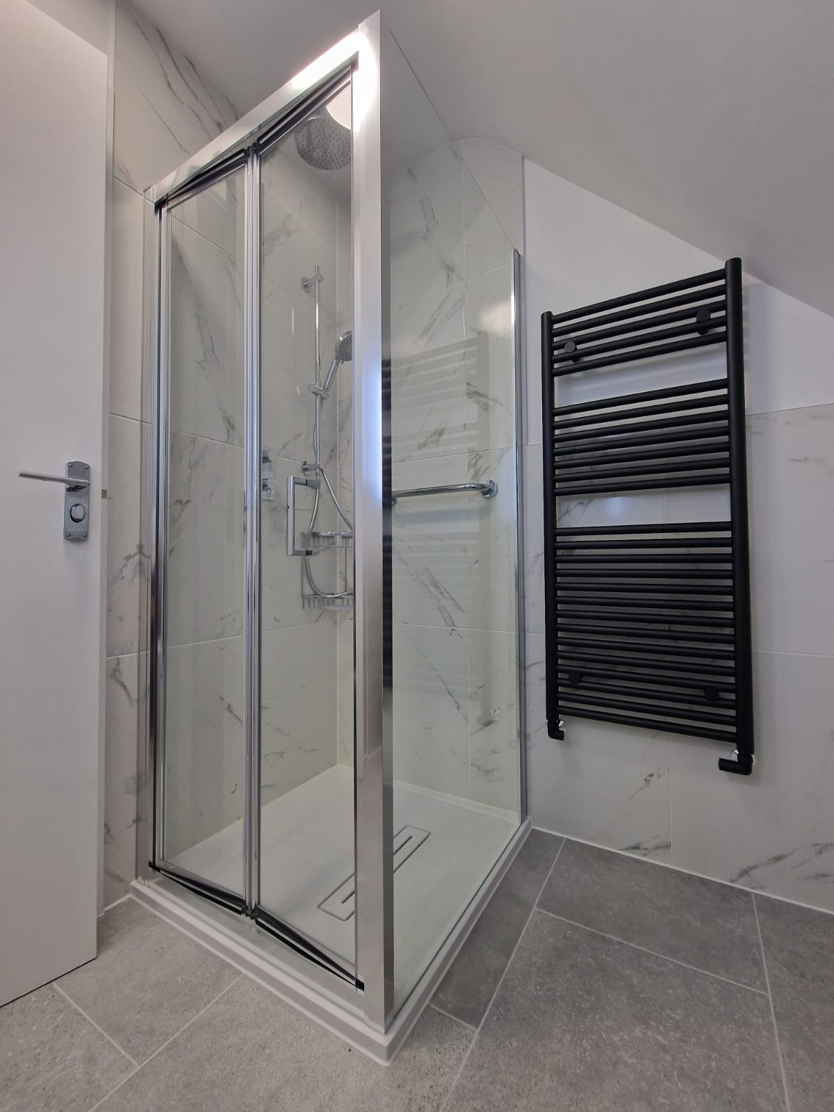 Nowoczesna kabina prysznicowa z przesuwnymi drzwiami, wykończona białymi płytkami imitującymi marmur, z czarnym grzejnikiem drabinkowym na ścianie i szarymi płytkami podłogowymi.