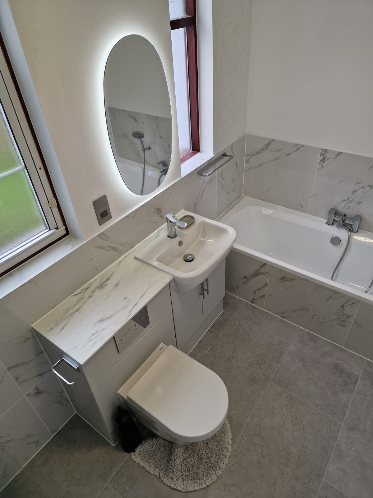 Nowoczesna łazienka z owalnym lustrem z podświetleniem LED, umywalką nablatową z szafką imitującą marmur, wanną i szarą podłogą z płytek.