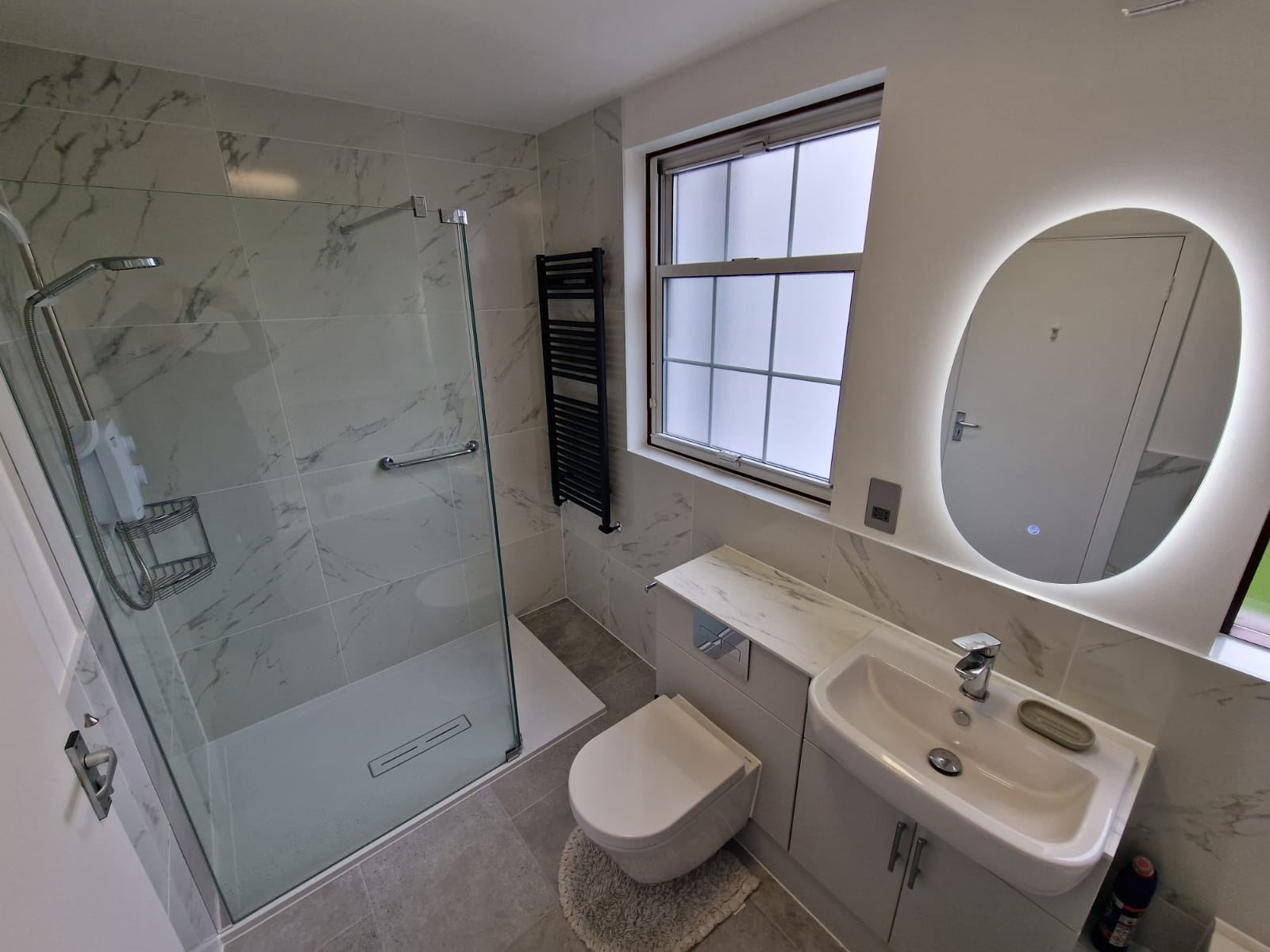 Nowoczesna łazienka z kabiną prysznicową typu walk-in, umywalką w zabudowie, podświetlanym lustrem owalnym i grzejnikiem drabinkowym w kolorze czarnym, wykończona płytkami imitującymi marmur.