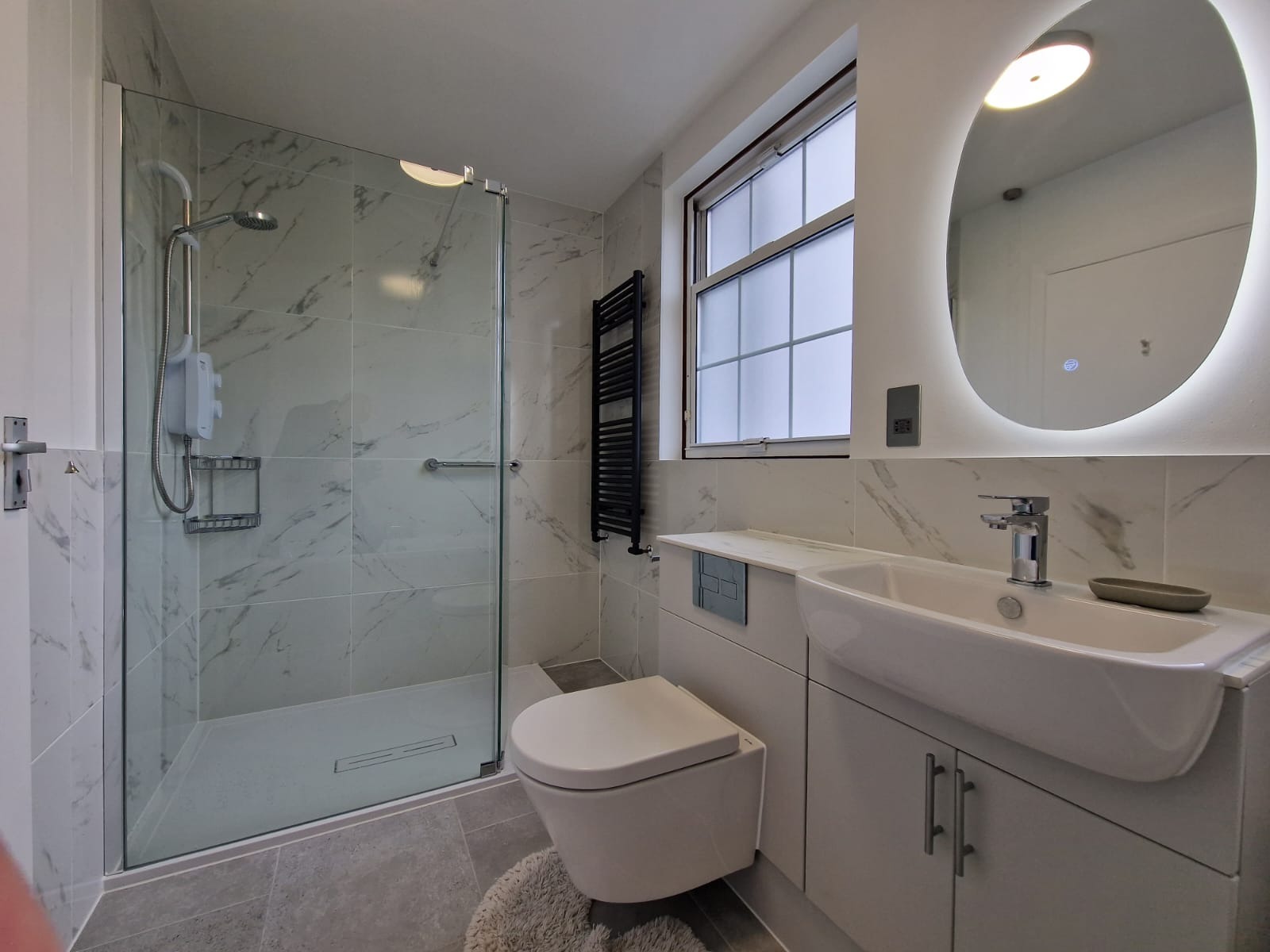 Nowoczesna łazienka w odcieniach szarości i bieli z kabiną prysznicową typu walk-in, podwieszaną toaletą i owalnym lustrem z podświetleniem LED.