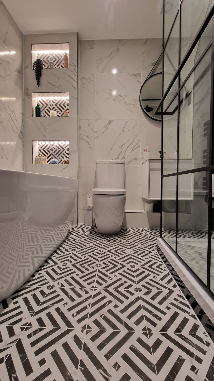 Nowoczesna łazienka w bieli i czerni z geometryczną posadzką, marmurowymi płytkami ściennymi, wanną, toaletą i kabiną prysznicową z czarnymi ramami.