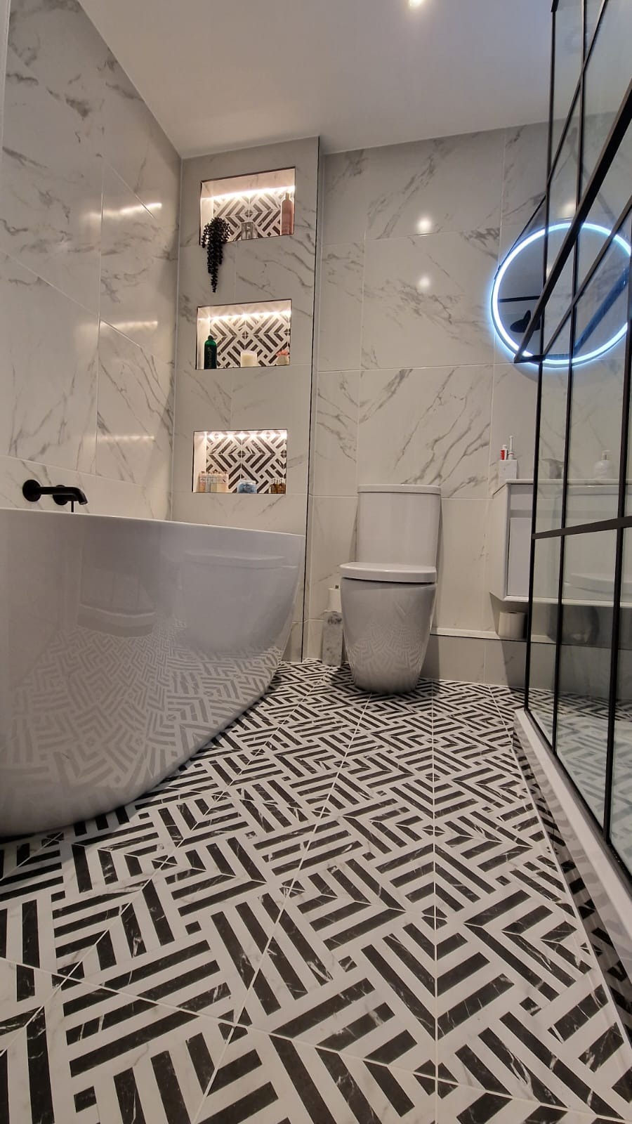 Nowoczesna łazienka z wolnostojącą wanną, toaletą, kabiną prysznicową z czarnymi ramami i geometryczną posadzką w czarno-białe wzory. W ścianie wnęki z podświetleniem i dekoracyjnymi elementami.