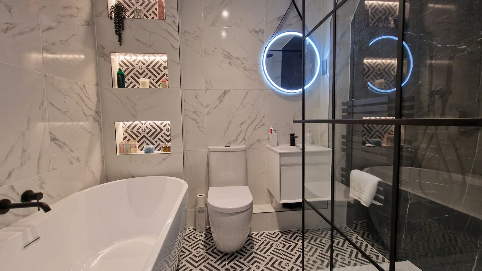 Nowoczesna łazienka z wanną wolnostojącą, toaletą kompaktową, umywalką nablatową i okrągłym lustrem z podświetleniem LED, wykończona płytkami imitującymi marmur i wzorzystą podłogą.