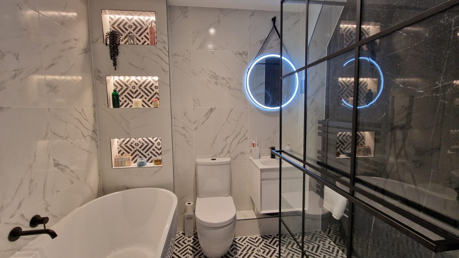 Nowoczesna łazienka z wanną wolnostojącą, toaletą, umywalką nablatową, kabiną prysznicową z czarnymi ramami, podświetlanym lustrem w kształcie okręgu i wnękami w ścianie z dekoracyjnym oświetleniem.