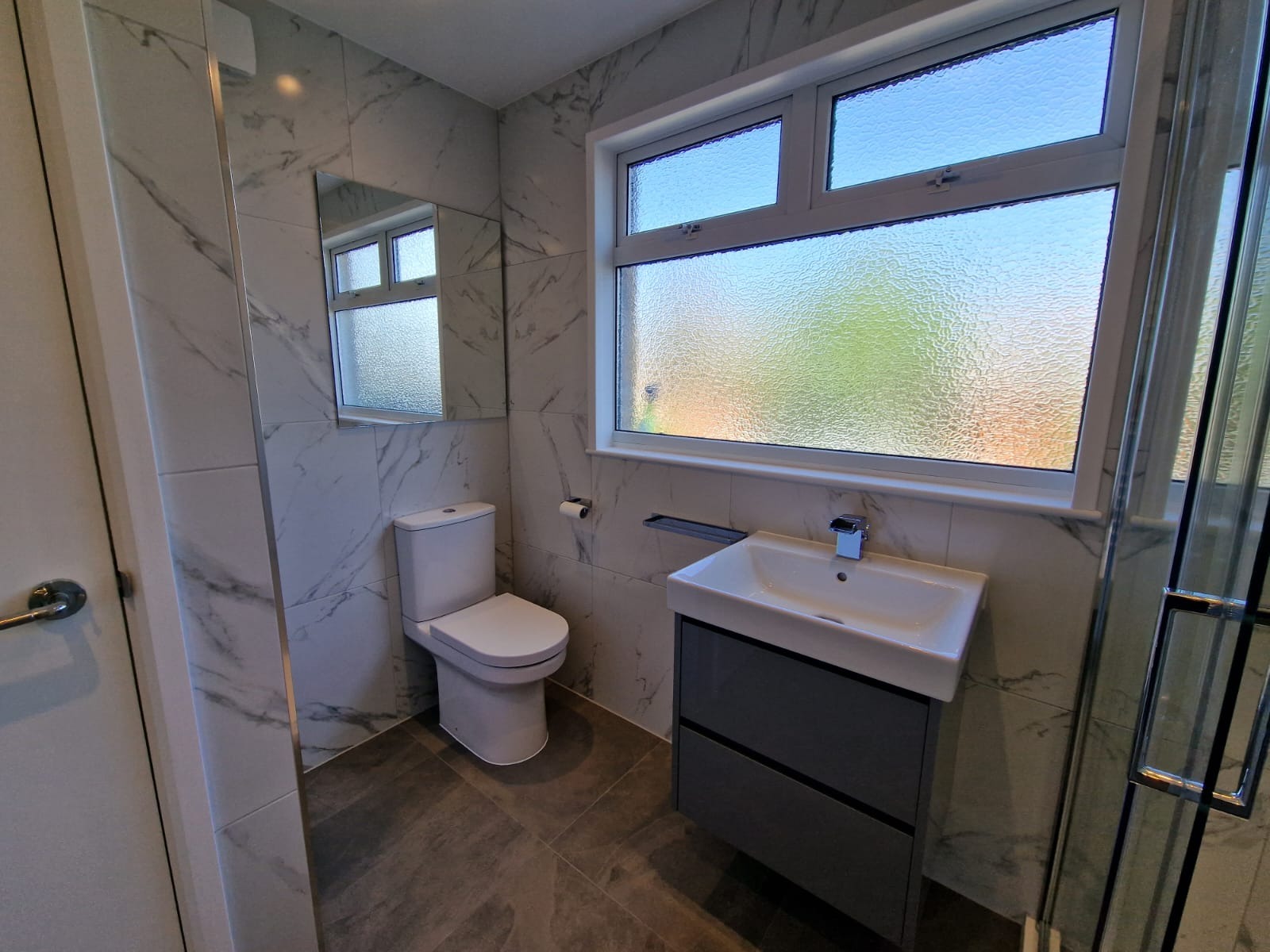 Nowoczesna łazienka z marmurowymi płytkami, wiszącą szafką pod umywalką w kolorze antracytowym, toaletą typu compact i oknem z szybą ornamentową