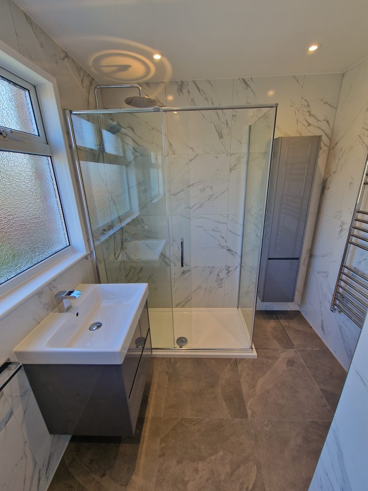 Nowoczesna łazienka z kabiną prysznicową typu walk-in, umywalką nablatową na szafce w kolorze grafitowym, grzejnikiem drabinkowym i płytkami imitującymi marmur na ścianach i podłodze.