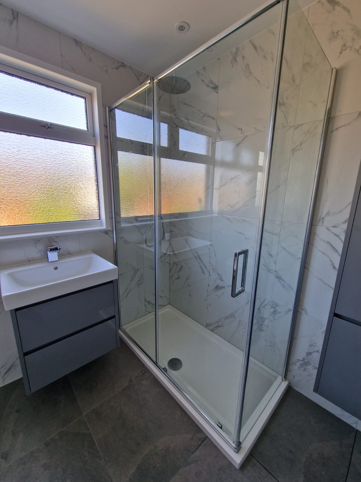Nowoczesna łazienka z kabiną prysznicową z deszczownicą, umywalką z szafką w kolorze szarym, marmurowe płytki na ścianach i okno z fakturą szkła.