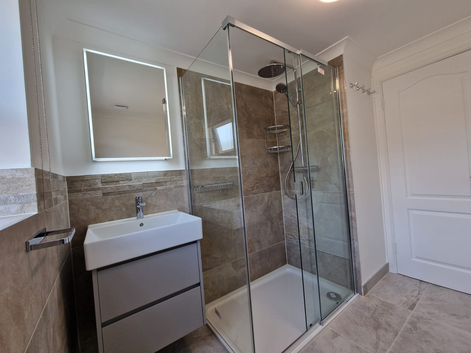 Wykończona łazienka z kabiną prysznicową typu walk-in, umywalką nablatową na szafce w kolorze szarym i lustrem z podświetleniem LED. Na ścianach płytki imitujące kamień w odcieniach brązu i beżu.