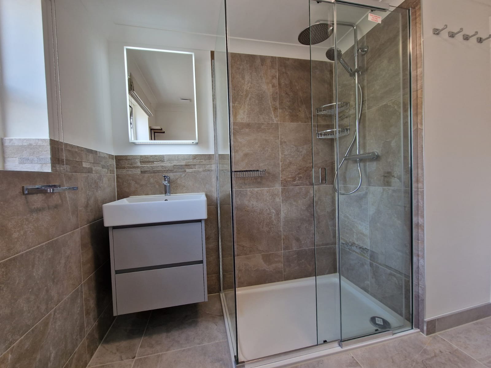 Nowoczesna łazienka z kabiną prysznicową typu walk-in, umywalką nablatową z szufladami w kolorze szarym, podświetlanym lustrem i brązowymi płytkami imitującymi kamień.