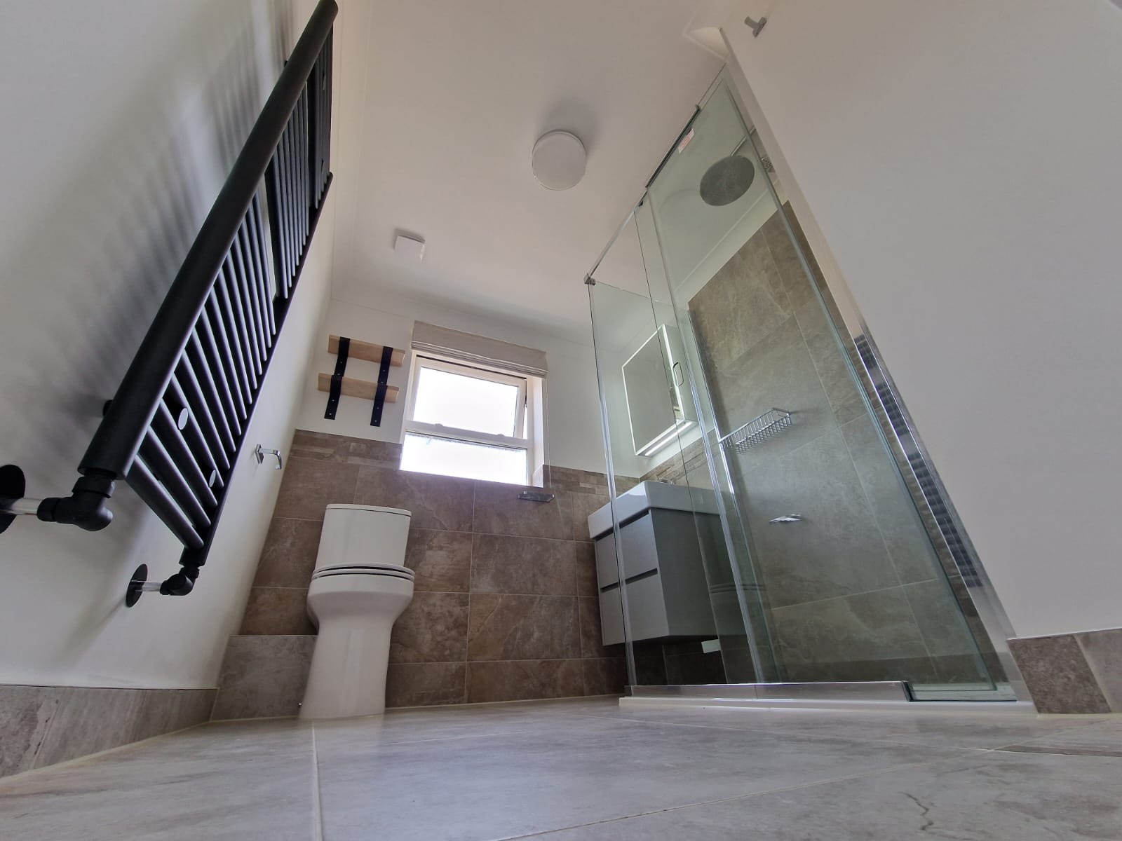 Nowoczesna łazienka z kabiną prysznicową ze szkła, grzejnikiem drabinkowym w kolorze czarnym i toaletą w jasnych barwach, widziana z perspektywy podłogi.