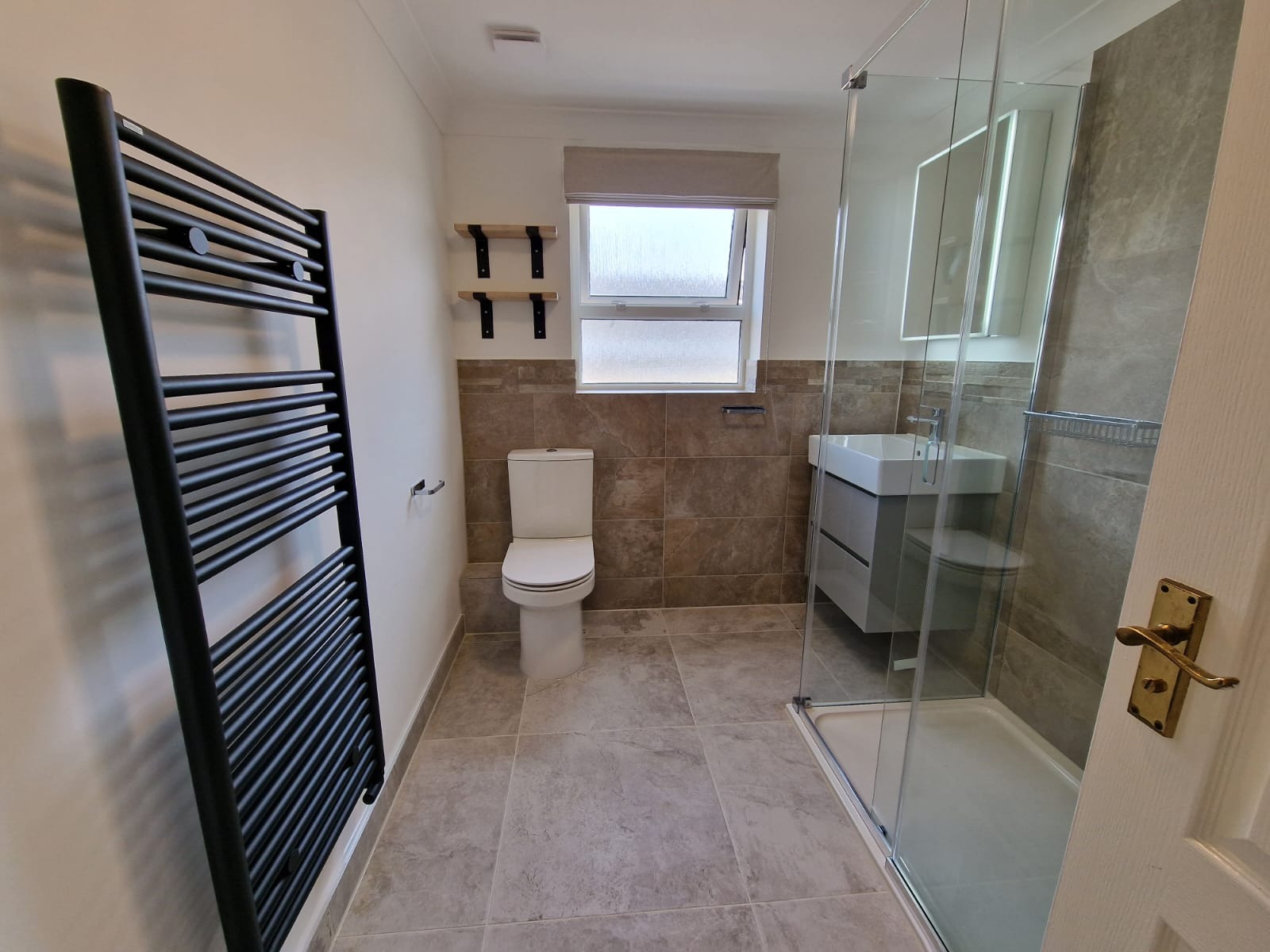 Nowoczesna łazienka z kabiną prysznicową, wiszącą szafką z umywalką, toaletą i czarnym grzejnikiem drabinkowym. Płytki w odcieniach beżu i szarości, drewniane półki nad toaletą.