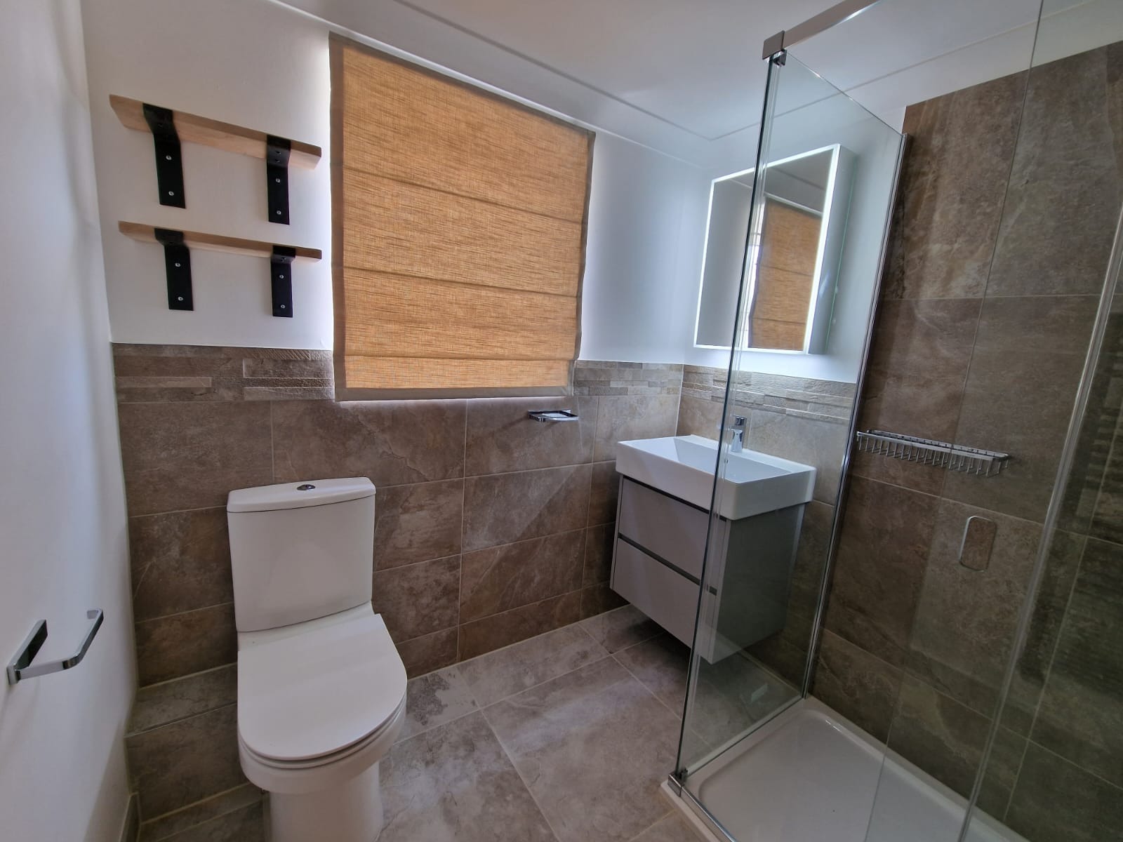 Nowoczesna łazienka z kabiną prysznicową ze szklaną ścianą, umywalką z szafką w odcieniach szarości, toaletą, brązowymi płytkami imitującymi kamień na ścianach i podłodze, oraz roletą rzymską...