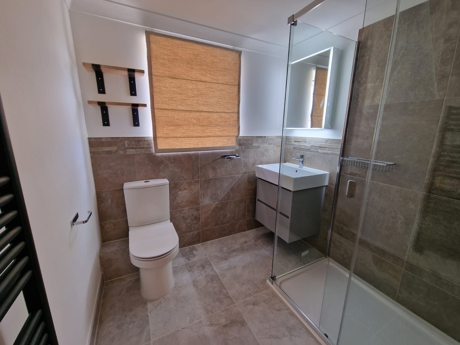 Wykończona łazienka z beżowymi płytkami, kabiną prysznicową ze szklaną ścianą, podwieszaną szafką z umywalką, toaletą i roletą rzymską w oknie.