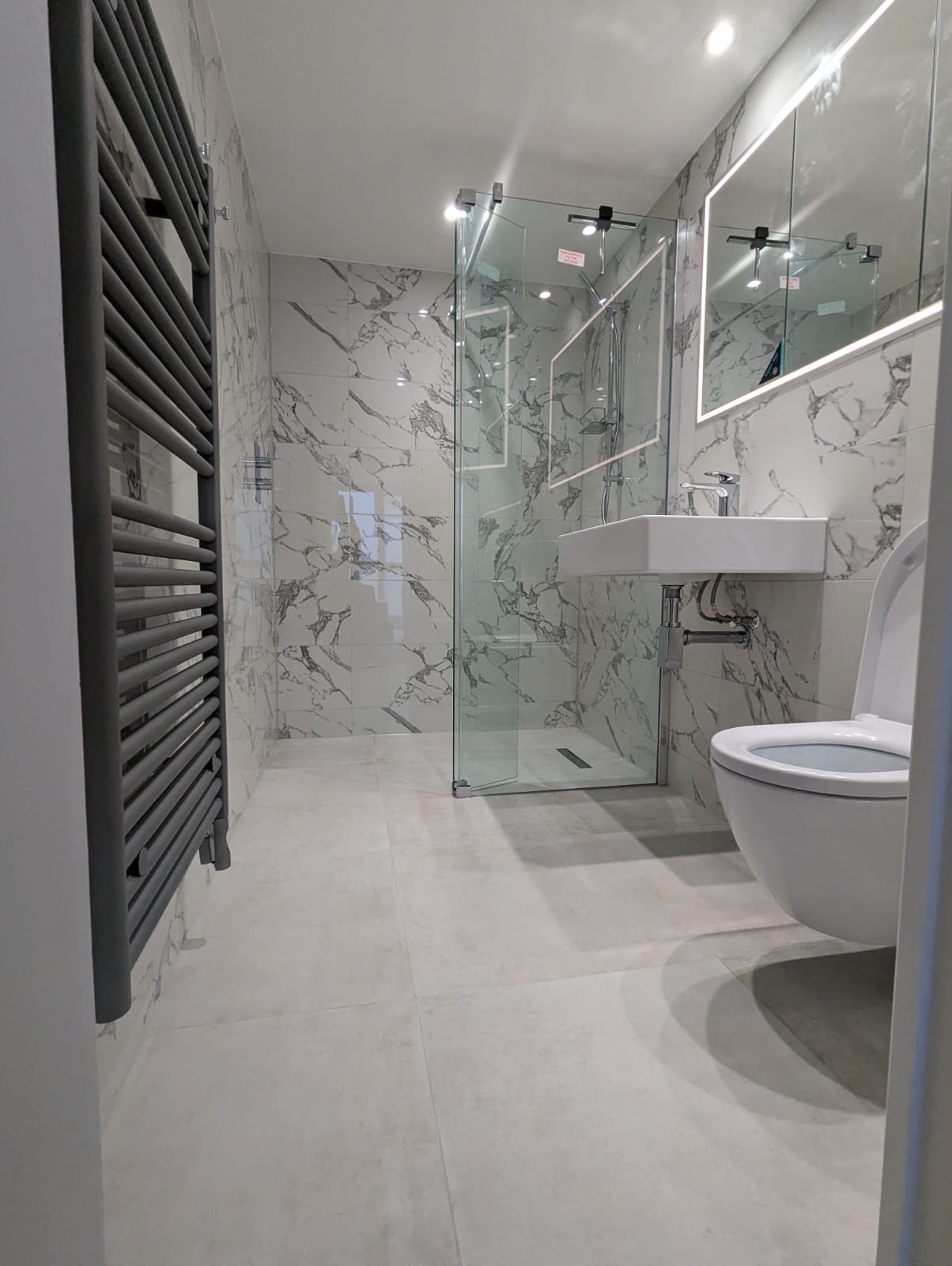 Nowoczesna łazienka z marmurowymi płytkami, przeszkloną kabiną prysznicową, podświetlanym lustrem i grzejnikiem drabinkowym w kolorze grafitowym.