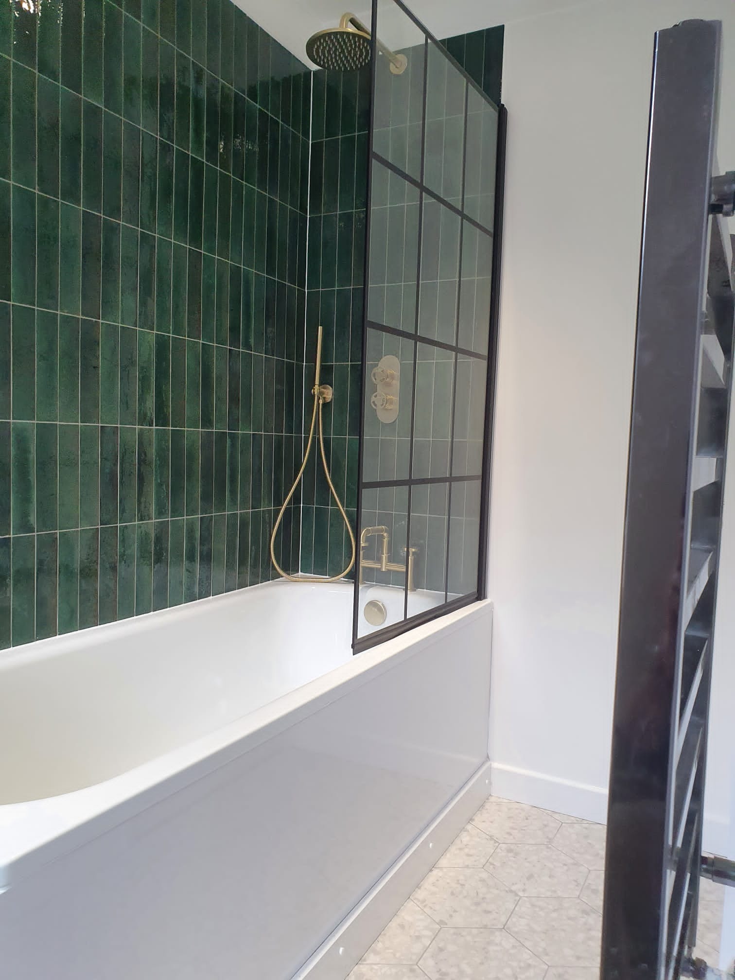 Biała wanna z zielonymi, pionowymi płytkami na ścianie, złota armatura i czarna szklana ścianka działowa z metalowymi szprosami.