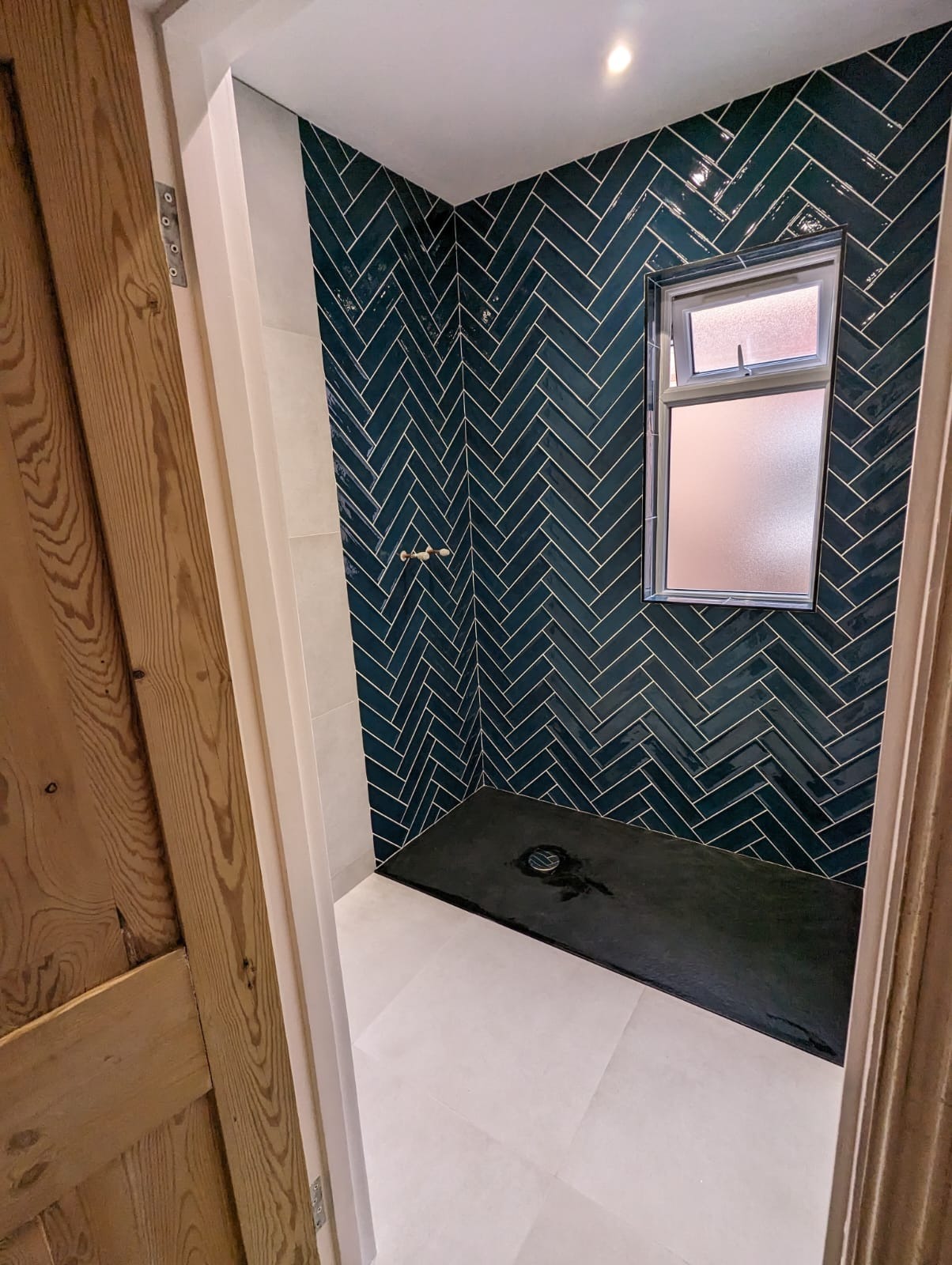 Wykończona kabina prysznicowa z ciemnozielonymi, błyszczącymi płytkami ułożonymi w jodełkę, małym oknem i czarnym brodzikiem, widok zza drewnianych drzwi.