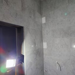 Termo Lux - Ściana wyłożona marmurowymi płytkami w łazience, widoczne otwory na instalacje i włączniki. W tle fragment oświetlenia budowlanego.