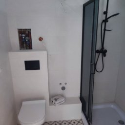 Termo Lux - Wykończona łazienka z białą zabudową toalety, czarną kabiną prysznicową i wzorzystą podłogą. Widoczna instalacja wodna i pudełko z akcesoriami.