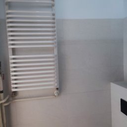 Termo Lux - Biały, drabinkowy grzejnik łazienkowy na jasnych, kwadratowych płytkach. Widoczne rury doprowadzające wodę i fragment szafki z czarnym elementem.