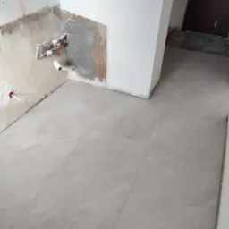 Jasne płytki podłogowe w trakcie remontu kuchni, widoczne instalacje wodne i fragment ściany z niedokończonym wykończeniem. Minimalistyczny styl, surowy stan.