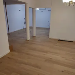 Jasna, drewniana podłoga ułożona w przestronnym wnętrzu z białymi ścianami i kolumnami. Widoczna drabina i narzędzie do montażu podłogi. Nowoczesny żyrandol sufitowy.