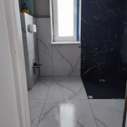 Wykończona łazienka z białymi i czarnymi płytkami imitującymi marmur, widoczne fragmenty instalacji i okno. Płytki ułożone z dbałością o detale.