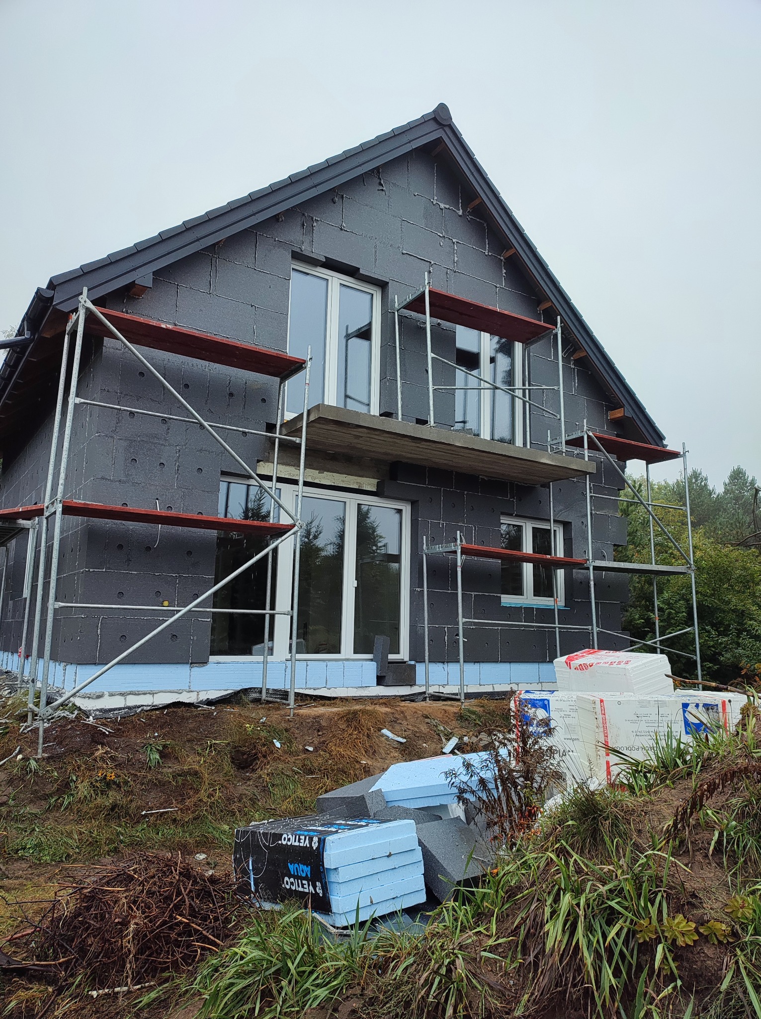 Dom jednorodzinny w trakcie ocieplania elewacji, widoczne rusztowania, płyty izolacyjne grafitowe i niebieskie styropianowe oraz materiały budowlane na ziemi.