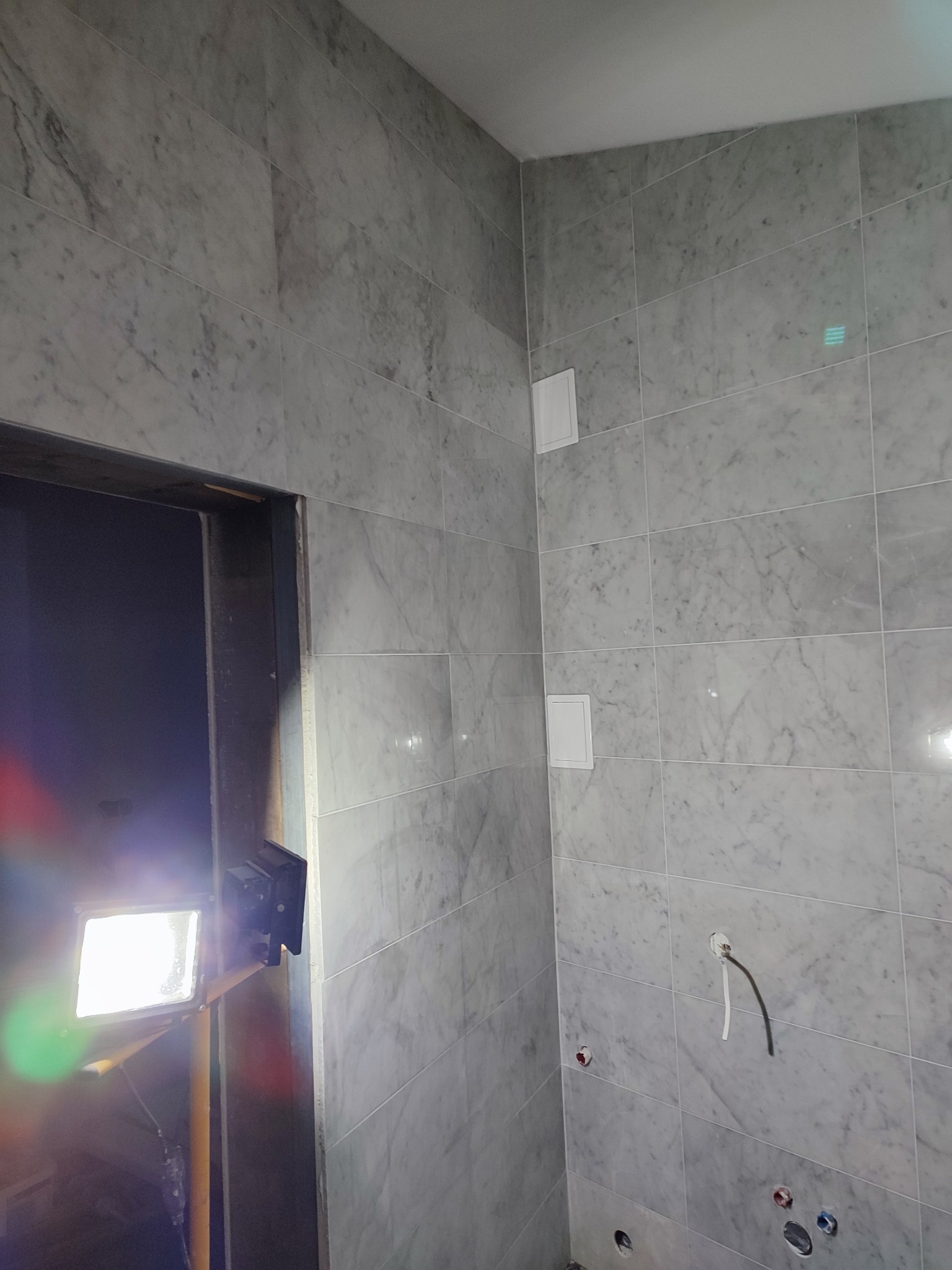 Ściana wyłożona marmurowymi płytkami w łazience, widoczne otwory na instalacje i włączniki. W tle fragment oświetlenia budowlanego.