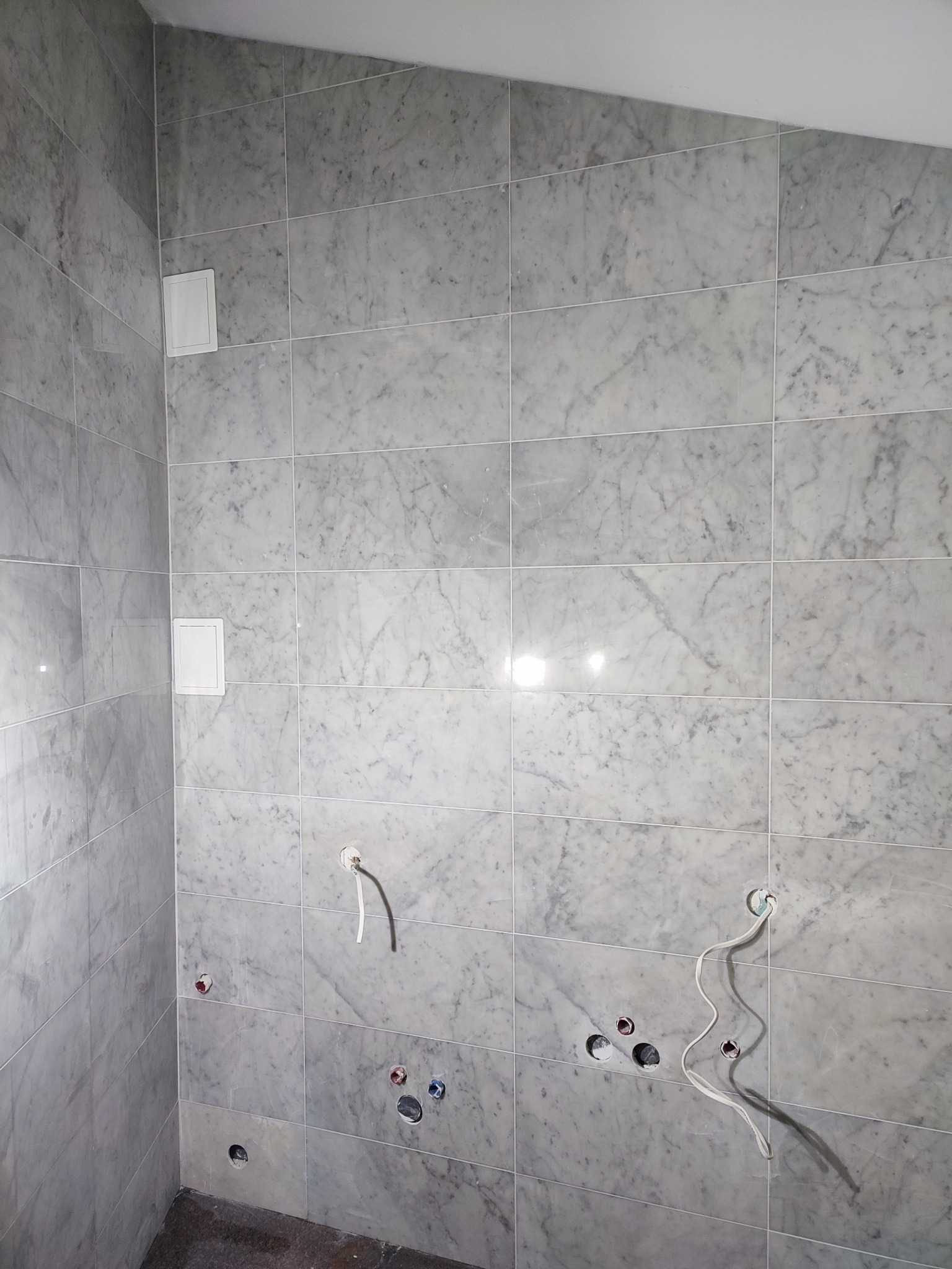 Wykończona łazienka z marmurowymi płytkami, widoczne otwory na armaturę i wystające przewody elektryczne, sugerujące etap prac instalacyjnych przed montażem wyposażenia.