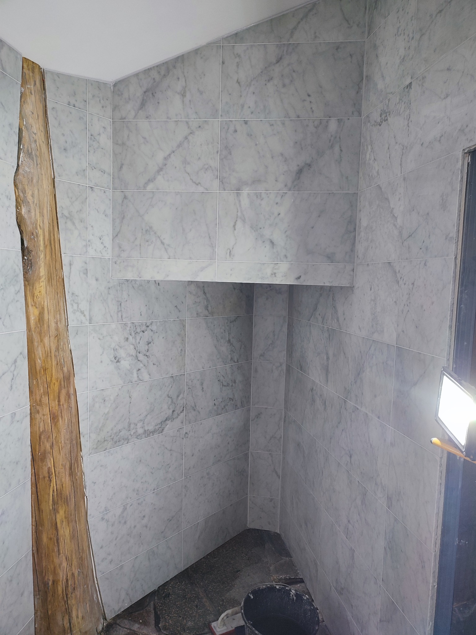 Wykończona łazienka z marmurowymi płytkami, wnęką i surowym drewnianym elementem. Wiadro na podłodze sugeruje trwające prace wykończeniowe.