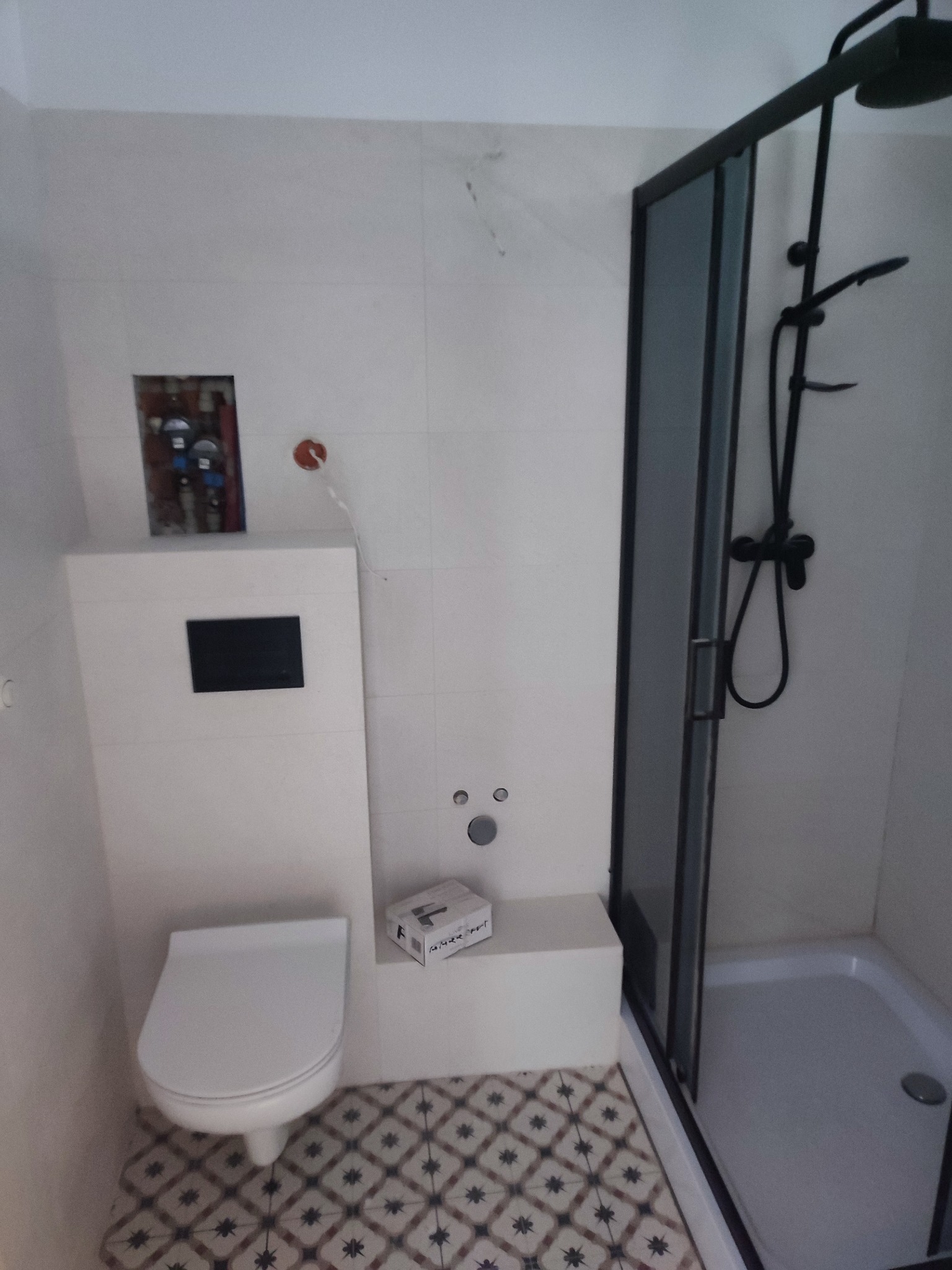 Wykończona łazienka z białą zabudową toalety, czarną kabiną prysznicową i wzorzystą podłogą. Widoczna instalacja wodna i pudełko z akcesoriami.