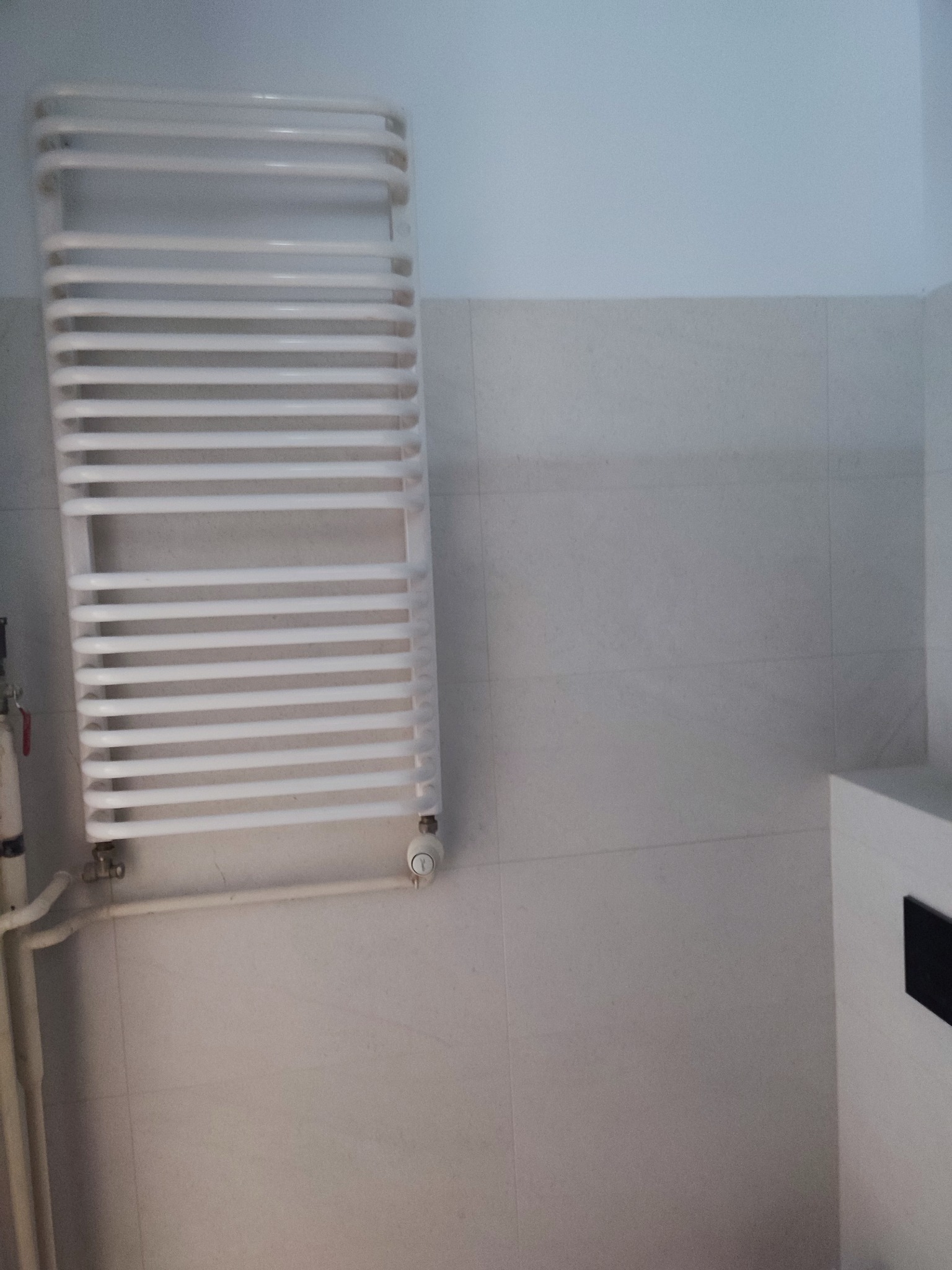 Biały, drabinkowy grzejnik łazienkowy na jasnych, kwadratowych płytkach. Widoczne rury doprowadzające wodę i fragment szafki z czarnym elementem.