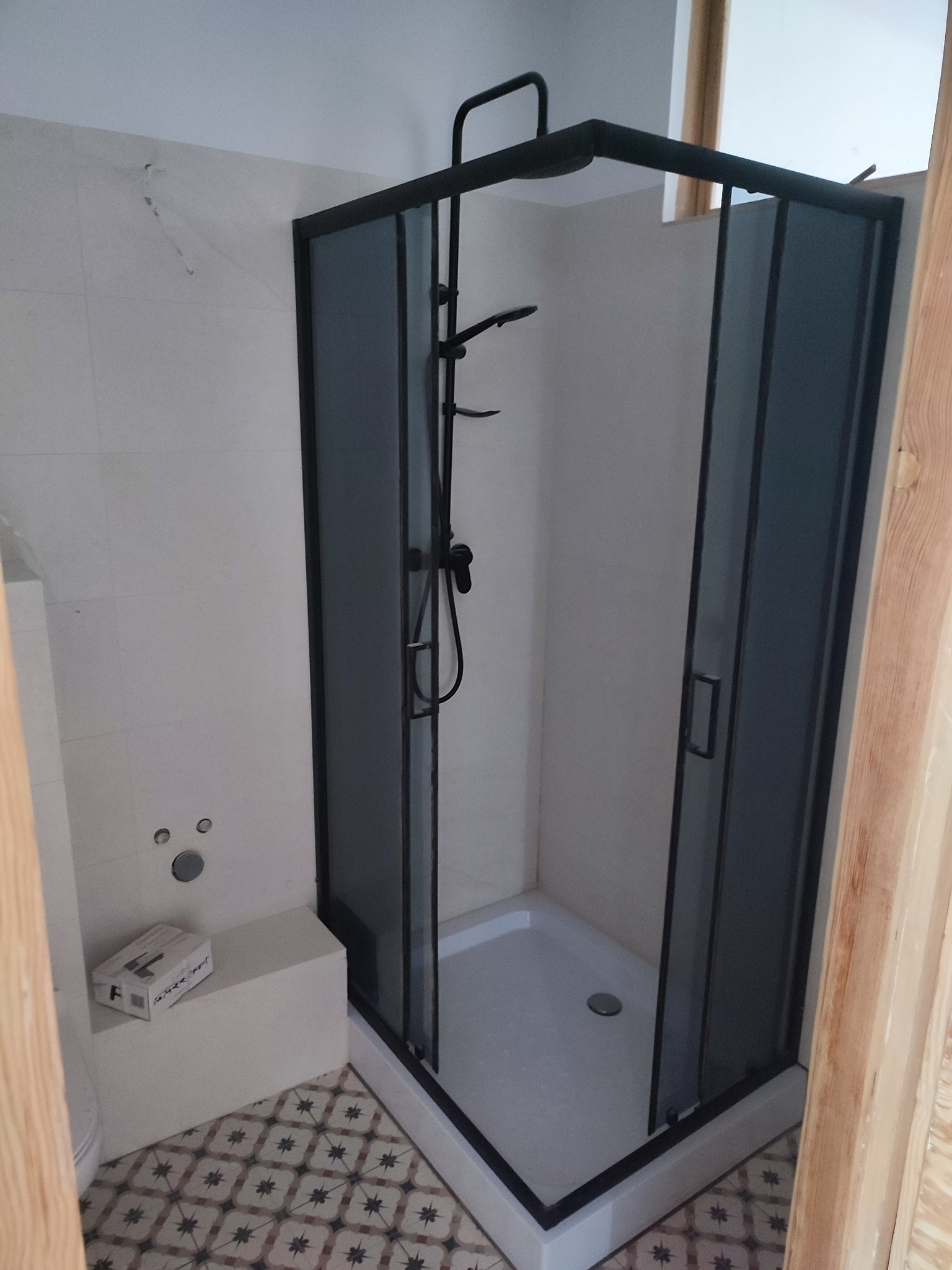 Nowoczesna kabina prysznicowa z czarnymi profilami i szklanymi ściankami, zamontowana w łazience z białą glazurą i wzorzystą podłogą.