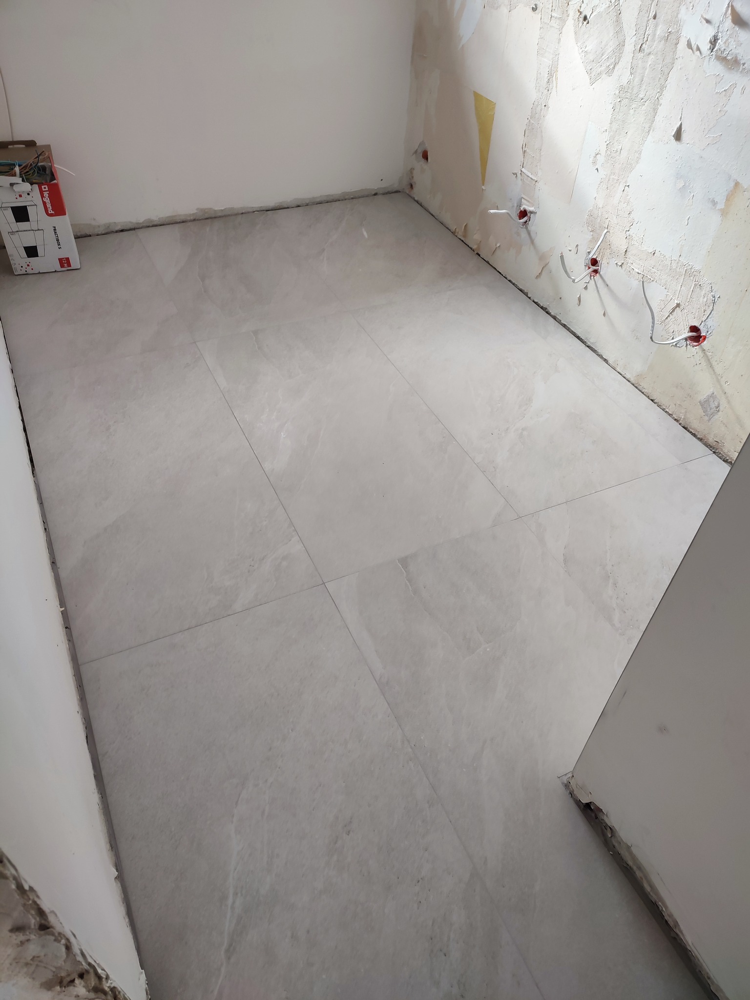 Ułożone jasnoszare płytki podłogowe w rogu pomieszczenia z surowymi ścianami i wystającymi przewodami elektrycznymi. Widoczna paczka płytek.