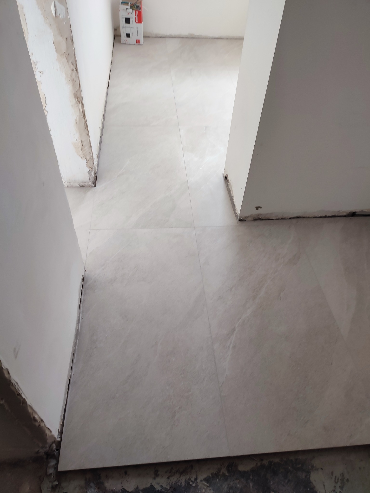 Jasne płytki podłogowe, ułożone w korytarzu podczas remontu. Widoczne ślady prac wykończeniowych na ścianach i podłożu. W tle kartonowe opakowania.