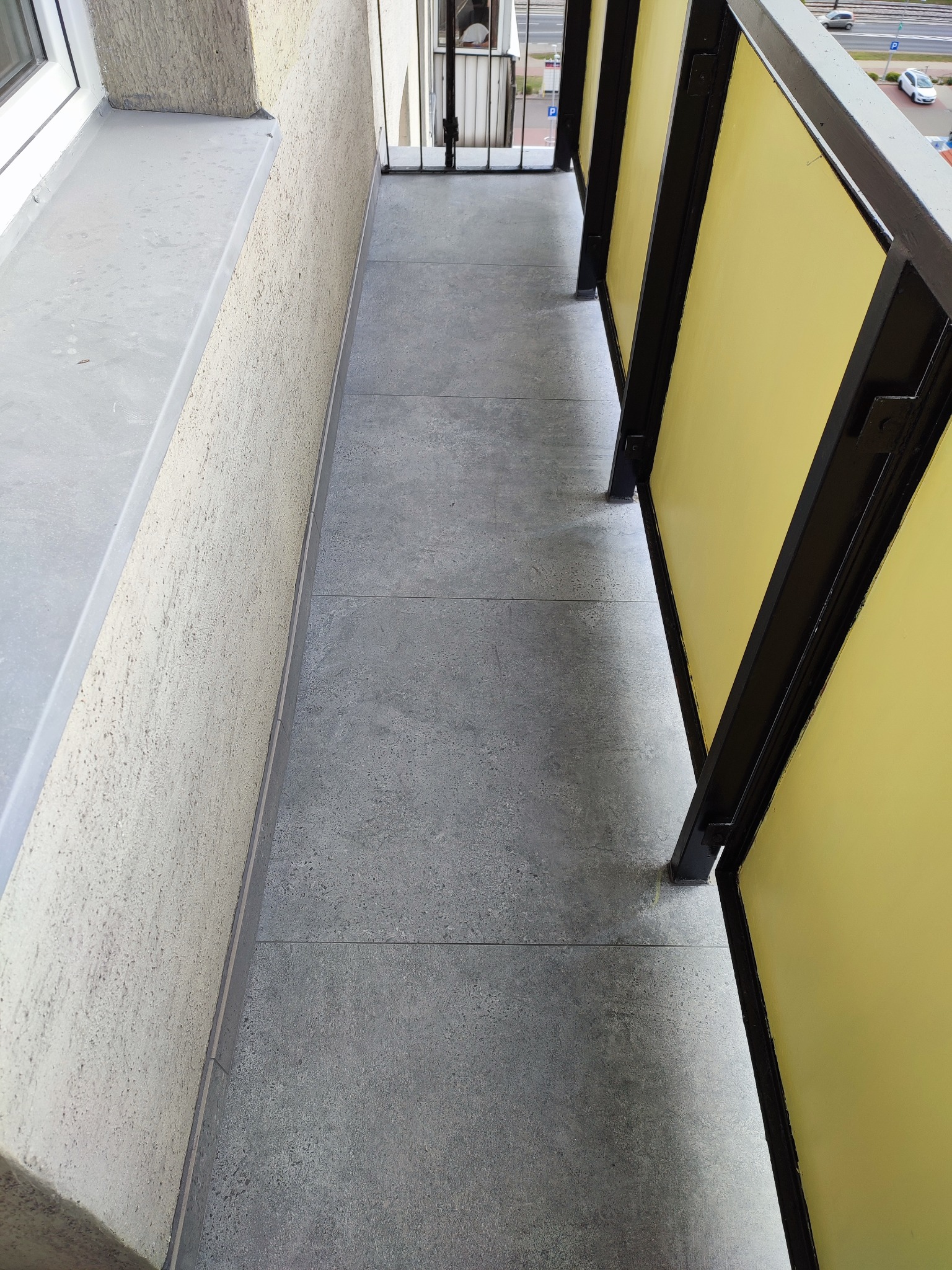 Wykończony balkon z szarymi płytkami podłogowymi i żółtymi panelami balkonowymi, widok z góry na zewnątrz budynku.