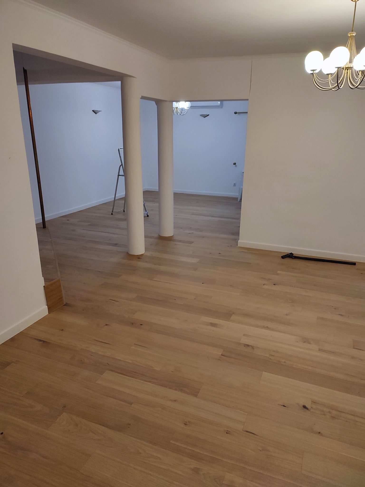 Jasna, drewniana podłoga ułożona w przestronnym wnętrzu z białymi ścianami i kolumnami. Widoczna drabina i narzędzie do montażu podłogi. Nowoczesny żyrandol sufitowy.