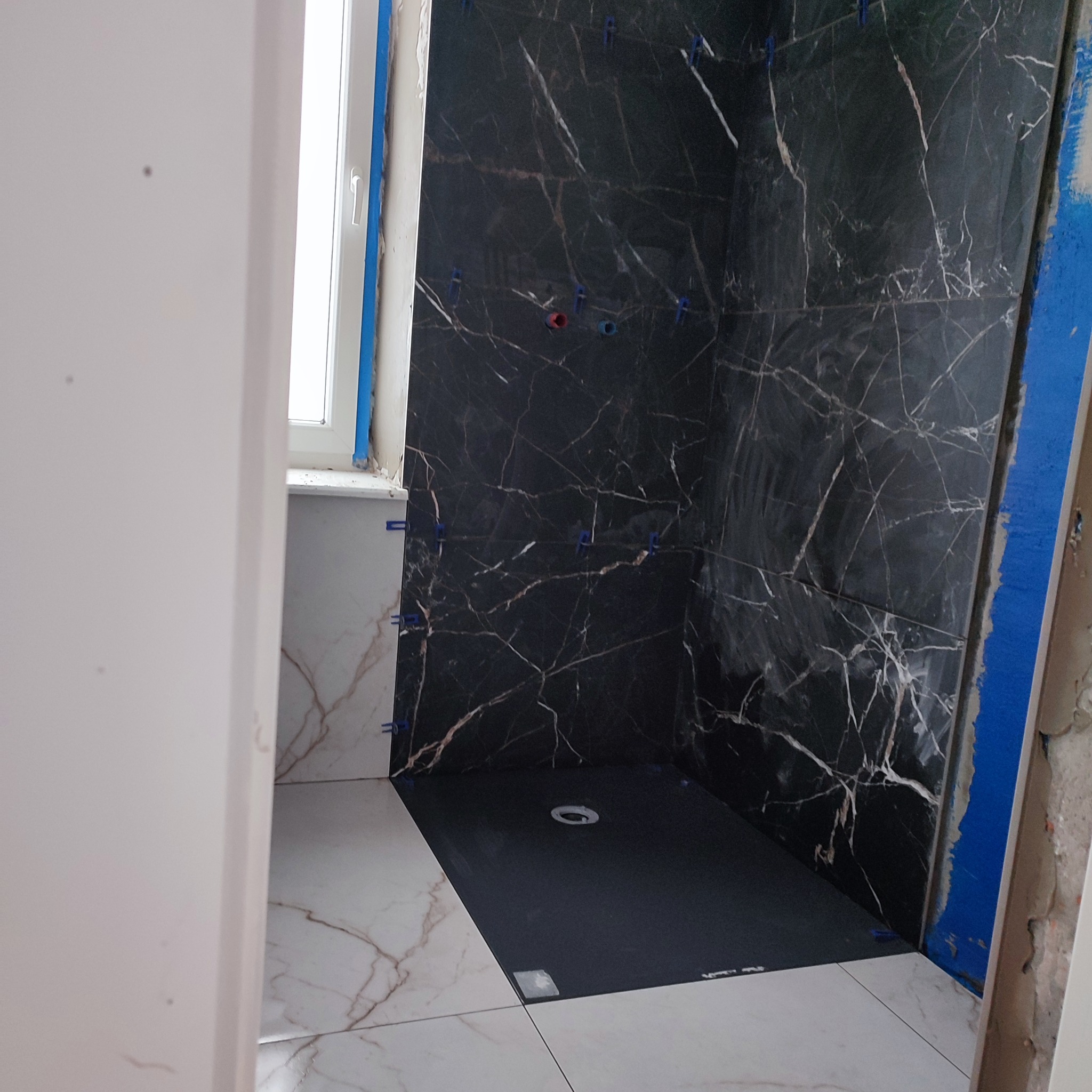 Wnętrze łazienki w trakcie wykańczania: czarne płytki imitujące marmur na ścianach kabiny prysznicowej, białe płytki podłogowe z brązowym wzorem.