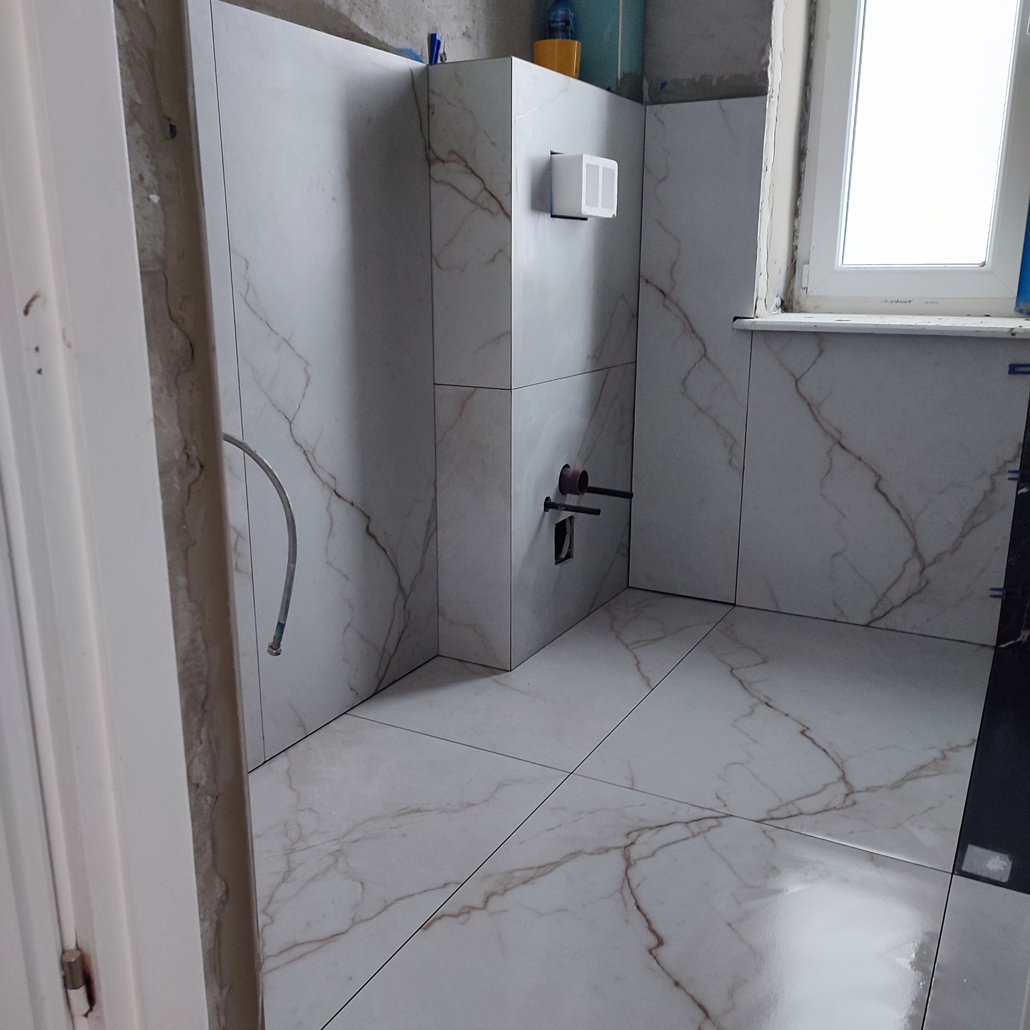 Ukończona łazienka w Krośniewicach: białe płytki imitujące marmur na ścianach i podłodze. Widoczne przyłącza wodne i okno. Estetyczne wykończenie.
