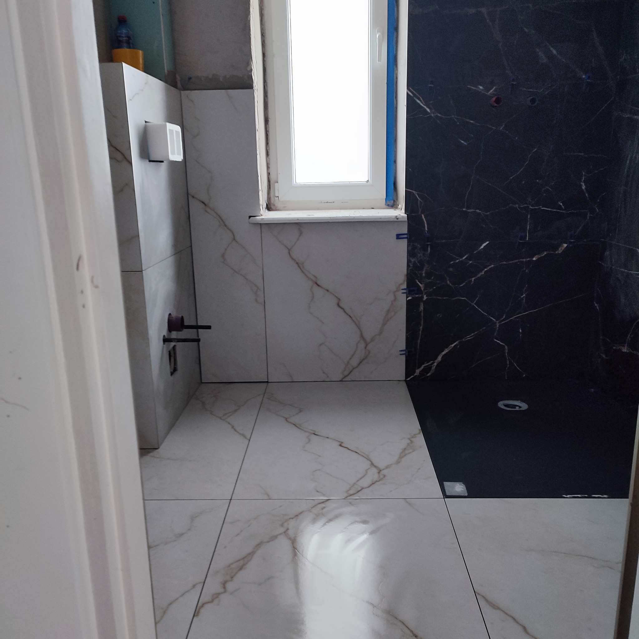 Wykończona łazienka z białymi i czarnymi płytkami imitującymi marmur, widoczne fragmenty instalacji i okno. Płytki ułożone z dbałością o detale.